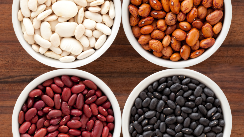 Four bowls of beans in various colors: white, brown, red, and black, set on a wooden surface, showcasing the protein-rich content in African Foods For Longer Hair.